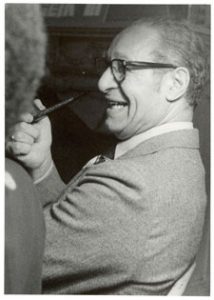 Black and white photo of Ted Berry seated and laughing with a pipe in his hand