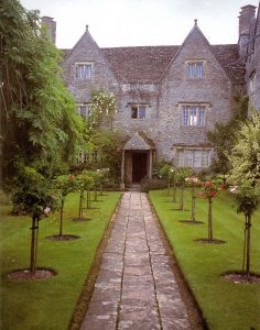 Color photo of the Kelmscott house, a large gray stone house with a large green lawn and a tree lined walkway
