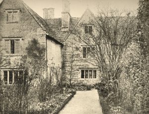 A black and white photograph of the rear of the manor house showing a gray stone estate with large chimmneys, lots of windows, and gardens