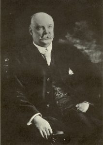 A black and white photograph of a bald older man with a white mustache seated
