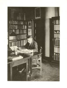 A man seated in a library-like study.  He is reading with bookshelves surrounding him.