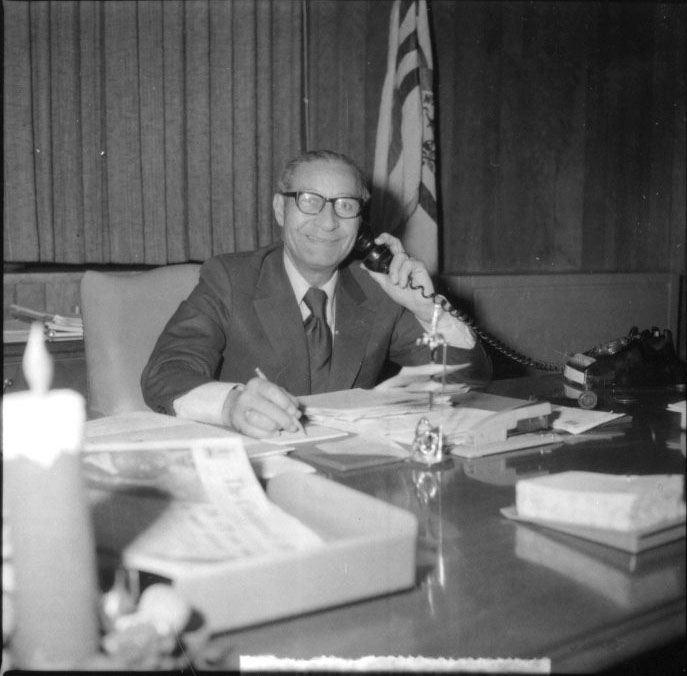 A black and white photograph of Theodore Berry sitting at his desk in a suit and glasses. He has a corded telephone up to his left ear and is smiling into the camera. He is holding a pen to a piece of paper in his right hand. 