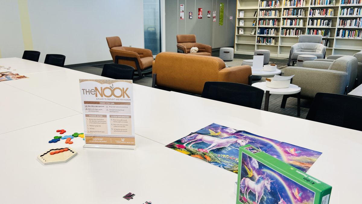 A long white table is topped with puzzles in progress and a sign in a signholder listing information about the new Nook area.