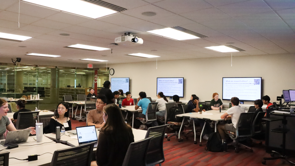 Classroom of students.  They sit in groups at tables that each have a computer monitor at the wall near them
