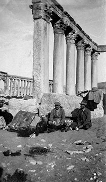 A black and white photo of two men seated in front of ruins. They are wearing hats and coats.
