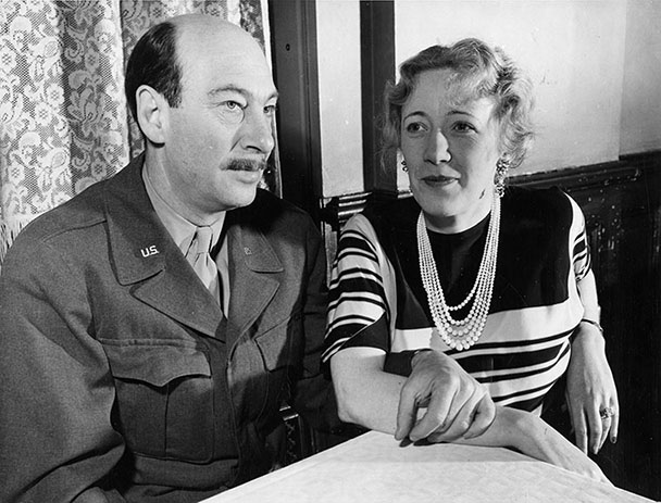A black and white photo of a man in a solider's uniform and a woman in a striped sweater. They are seated and linking arms.