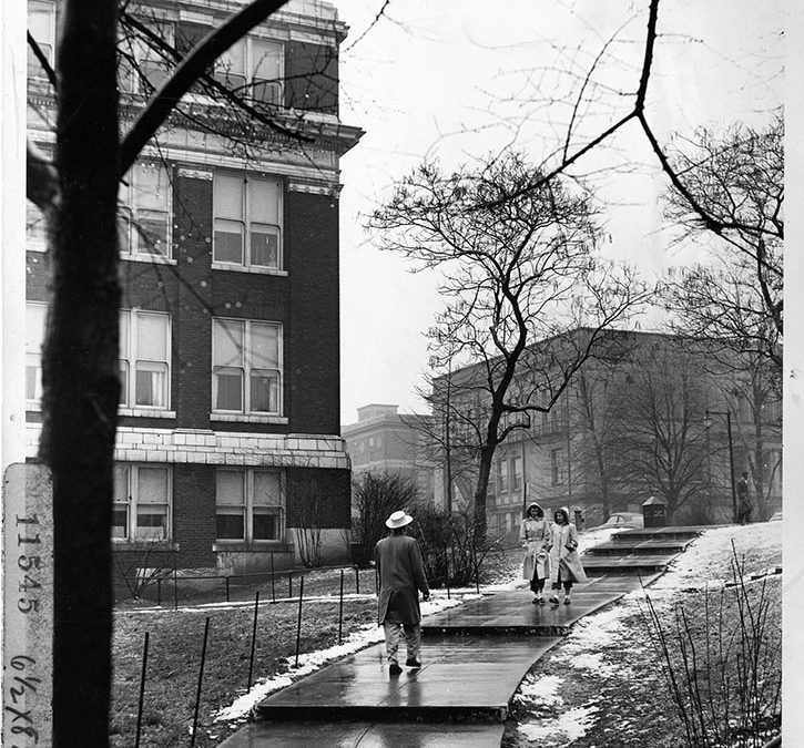 A black and white photo showing three people walking up an outdoor stairway in the snow surrounded by university buildings and trees.