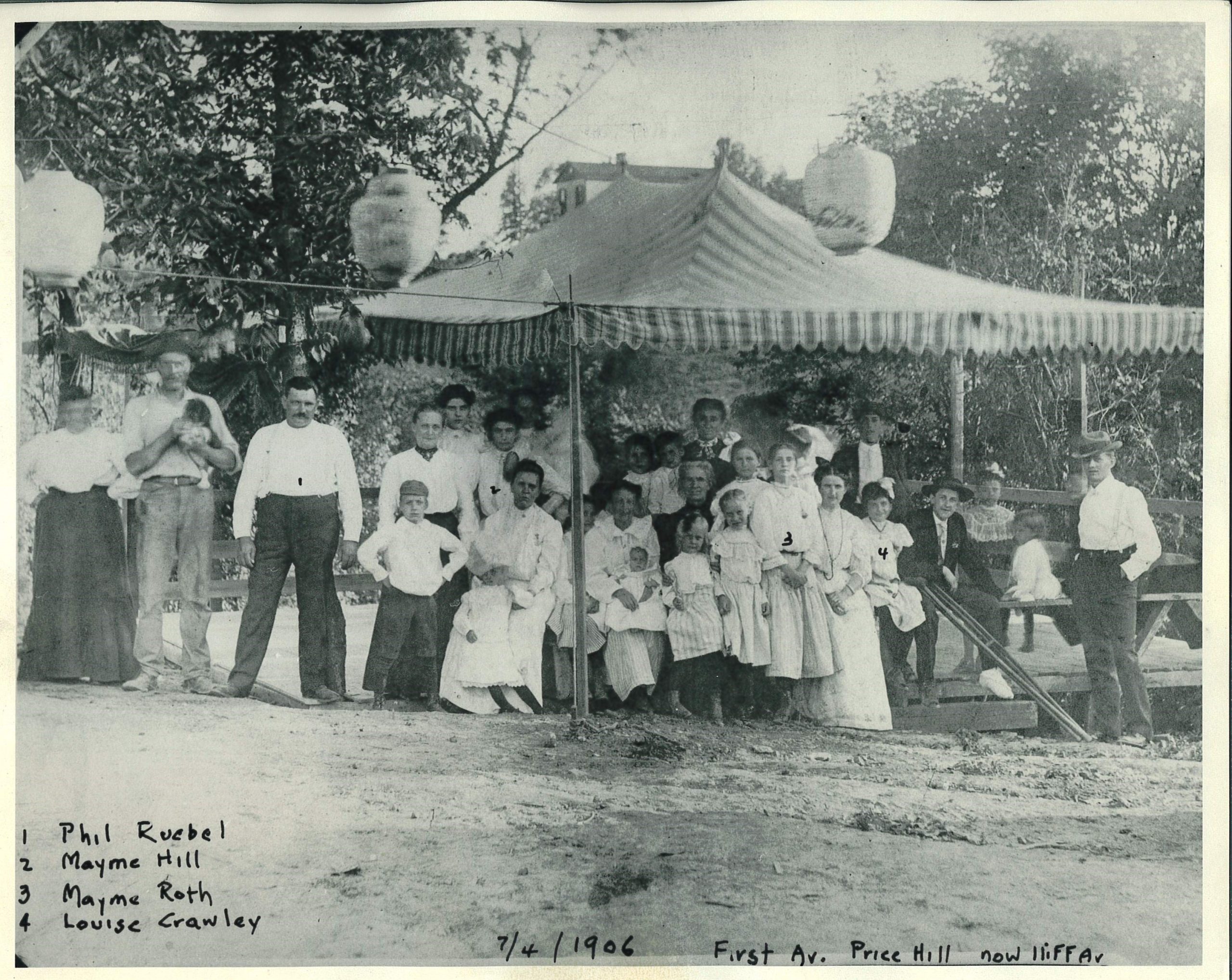 A group of 20-25 people pose for a group photograph under a decorated gazebo at a park outing. 