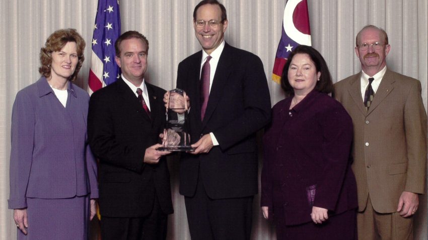 Governor Bob Taft stands in the center of photo holding award with two UC staff on either side of him smiling for photo in front of an American flag and Ohio state flag.