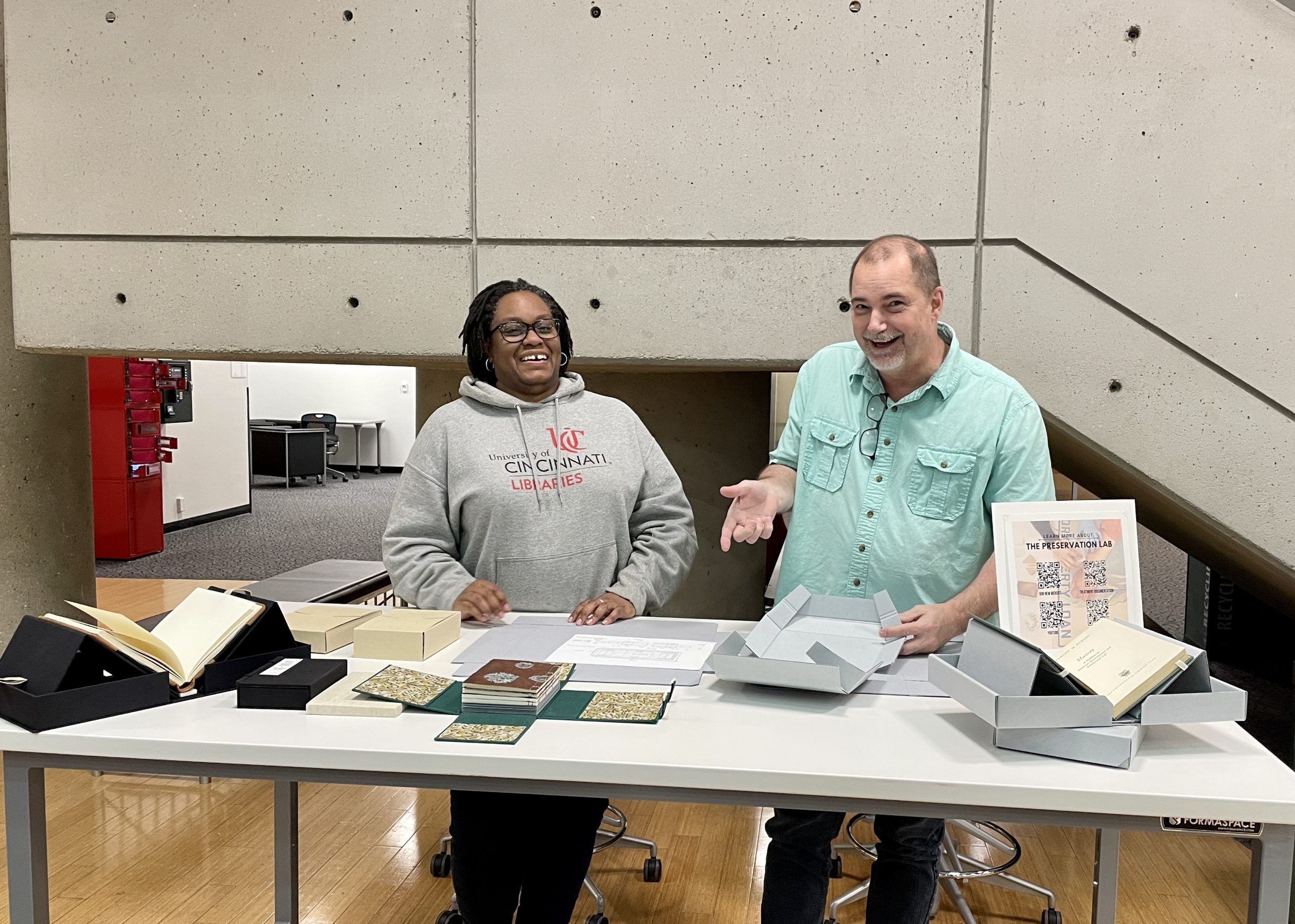 Chris and Hyacinth behind table that displays various enclosures to protect books.  Enclosures are made of binder board, corrugated blue board, and book clothe.  