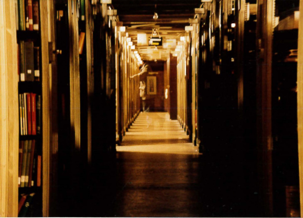 Creepy image of library stacks with a shadowy, half exposed man walking across the stacks