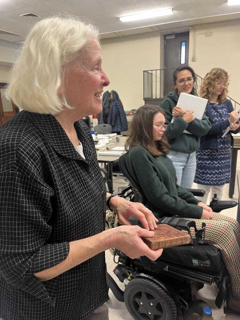 Julia Miller is in the fore ground holding a highly decorated book that is read with gold stamping and tooling. 