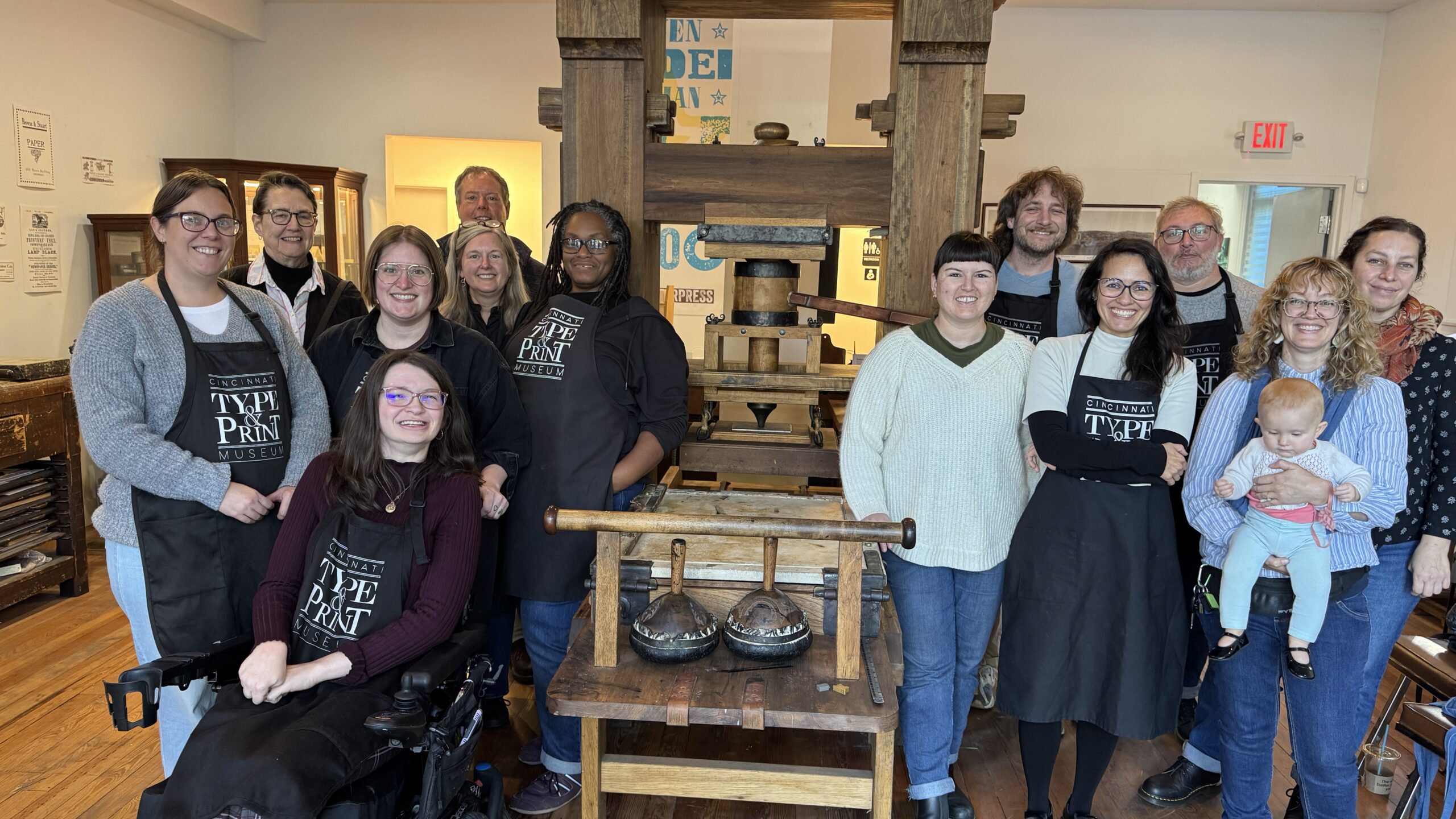 13 people and a baby posing in front of a large wooden printing press.