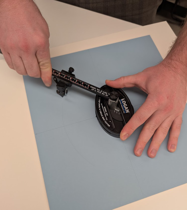 Two hands hold a compass-like mat cutter to cut a circle in a blue piece of mat board.