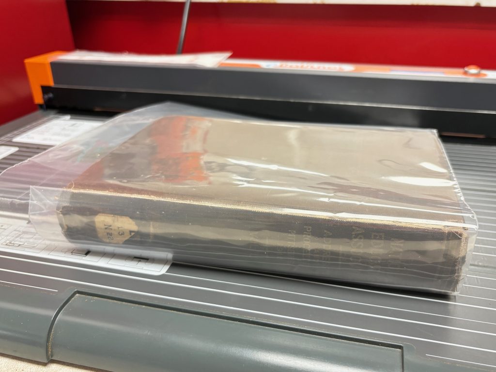 Book with jacket and spine pocket welded at the top edge.  The un-welded bottom of the jacket and pocket protrudes out from below the book in excess.