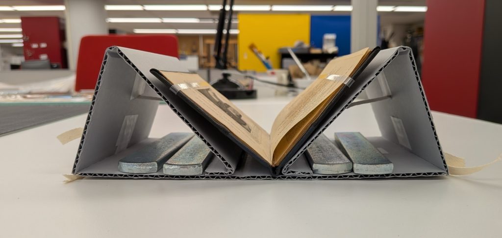 View of a small book strapped in a corrugated cradle with weights in along the base to hold the cradle in place.
