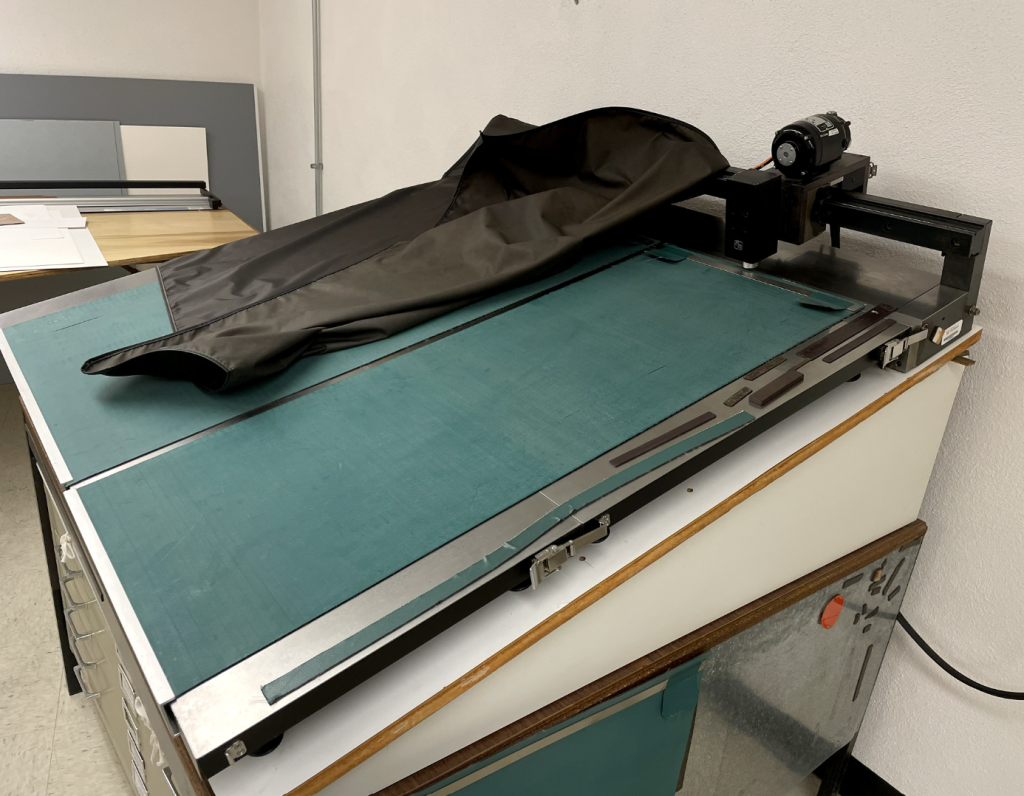 Metal table that's on a tilt that's partially uncovered below a black cover.  Green magnetic blankets cover table. At top of table is an arm with a motor, converter and anvil that slides across the top of the table.