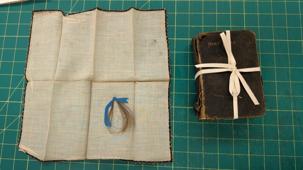 Unfolded handkerchief with a small lock of hair tied with a blue ribbon, sitting next to the cloth bound bible, tied with cloth string.
