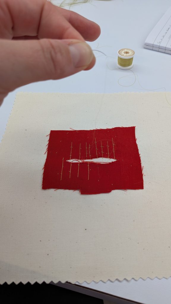 Red fabric with tear through the middle being repaired with needle and thread.